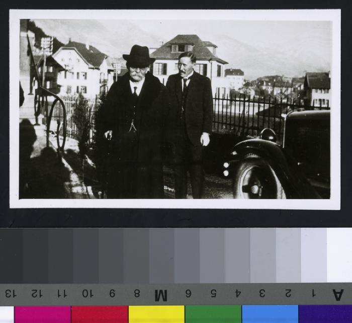 Portrait en pied du Prof. Dr. César Roux et d'un personnage. A droite, une voiture. Au fond des maisons et les montagnes.