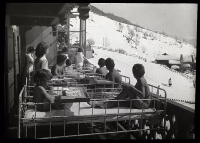 Enfants allités en cure de soleil et d'air pur sur une véranda. Des infirmières prennent soin d'eux.