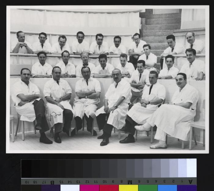 Portrait du Professeur Pierre Decker (3e au premier rang, à partir de la droite) entouré d'un groupe de jeunes médecins. Ils sont dans le grand auditoire de l'annexe chirurgicale de l'Hôpital cantonal.