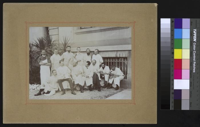 Portrait d'un groupe de médecins et d'assistants sur la cour de l'Hôpital cantonal. Le docteur Paul Narbel, assis au premier rang, bras levé, tenant un couteau à la main porte une drôle de chapeau conique sur la tête.