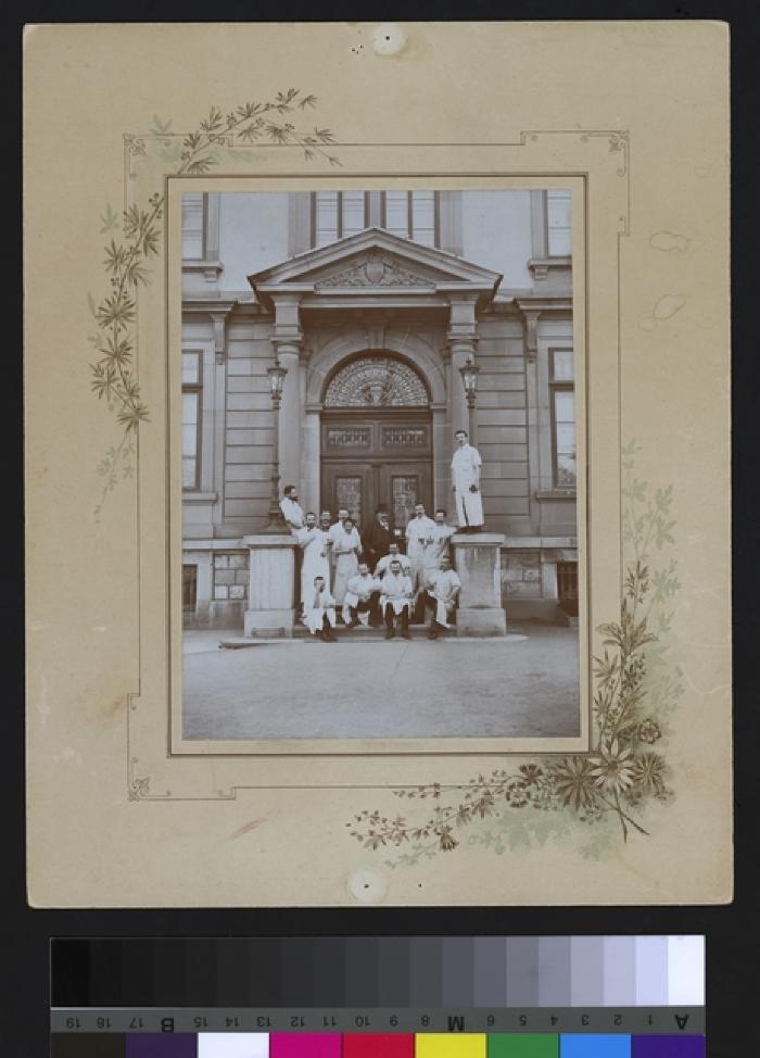 Portrait d'un groupe de médecins et d'assistants devant l'entrée principal de l'Hôpital cantonal. Le docteur Paul Narbel est débout, au deuxième rang, appuyé contre le muret, une pipe à la bouche.