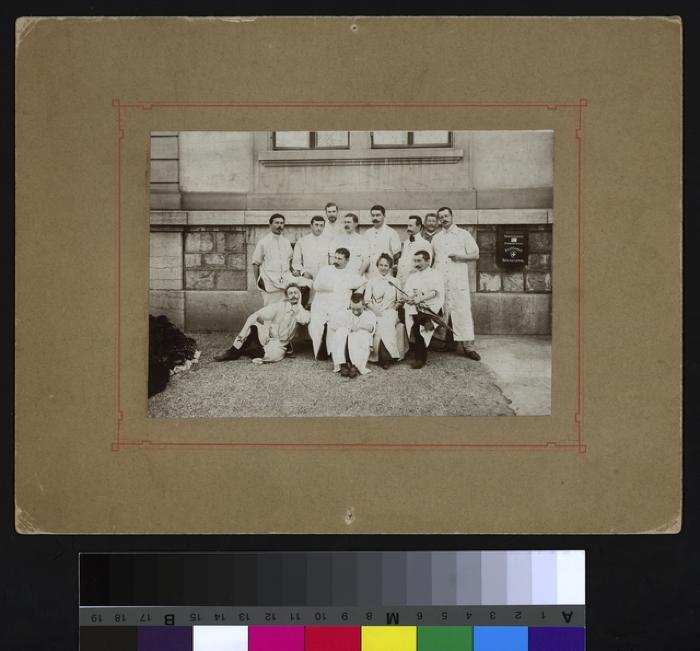 Portrait d'un groupe d'assistants, composé de douze hommes et d'une femme, posant en uniforme (blouse blanche de médecin). A l'arrière, une partie de la façade de l'hôpital cantonal et une boîte à lettres. Le Dr Francis Cevey est le cinquième à partir …