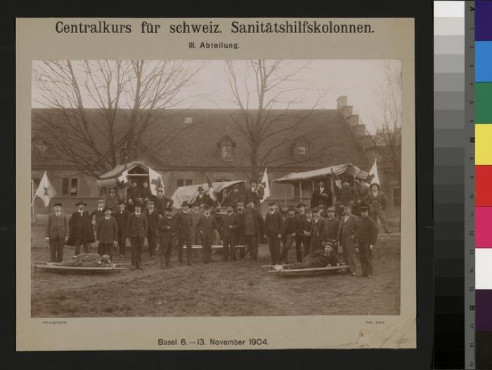 Portraits des participants par équipes au cours d'aides sanitaires qui eut lieu à Bâle du 6 au 13 novembre 1904. Les portraiturés sont debout, assis sur des charrettes ou couchés sur des brancards sur un champ devant une grande bâtisse .