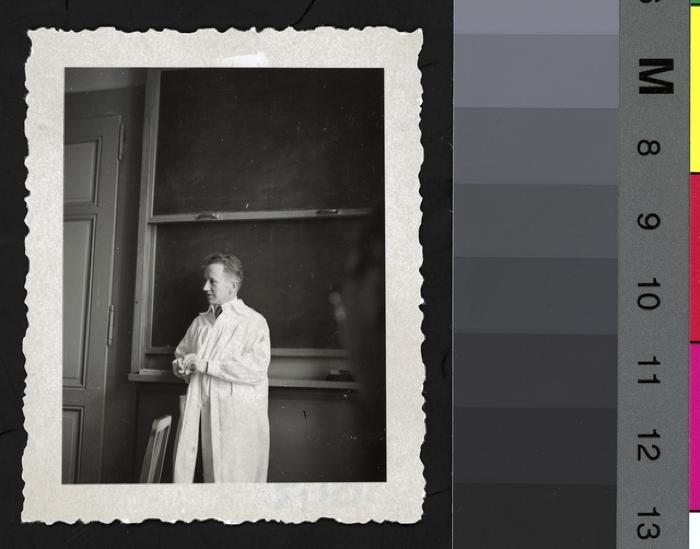 Portrait en pied du Prof. Amsler Marc dans une salle de cours, habillé avec une blouse blanche de médecin. Derrière lui, le tableau noir.