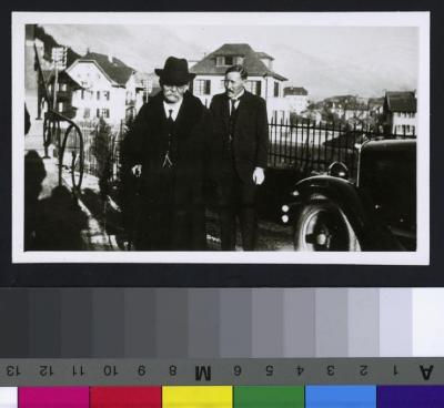 Portrait en pied du Prof. Dr. César Roux et d'un personnage. A droite, une voiture. Au fond des maisons et les montagnes.