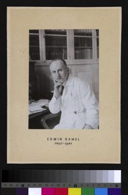 Portrait assis du professeur Edwin Ramel. Il est probablement dans un bureau car il y a une table de travail à droite de l'image et au fond une armoire avec des livres. Il porte la blouse de médecin.