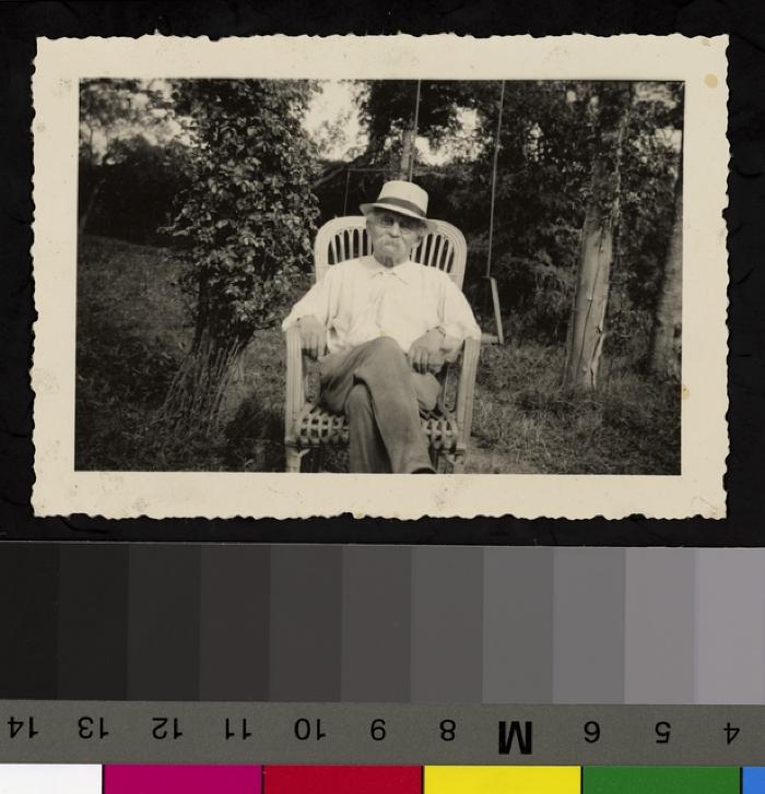 Portrait assis du Prof. Dr. César Roux âgé, posant sur une chaise au milieu d'un jardin. Il porte des habits légers et un chapeau de couleur claire. Derrière la chaise, une balançoire.