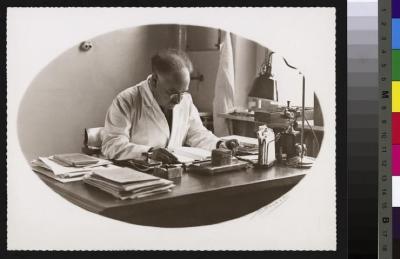 Portrait du Dr. Alfred Rosselet, assis à sa table de travail, laquelle est recouverte d'objets divers: feuilles, presse papier, papiers, lampe, téléphone. Le médecin a une cigarette dans la main gauche. A droite de l'image, une fenêtre avec un rebord s…