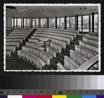 Intérieur du grand auditoire de la nouvelle Ecole de Médecine. Vue de l'amphithéâtre, ses tables et ses chaises ainsi que de la paroi vitrée depuis laquelle on aperçoit, en arrière plan, le quartier de la cité et de la cathédrale. Le plafond de l'audit…