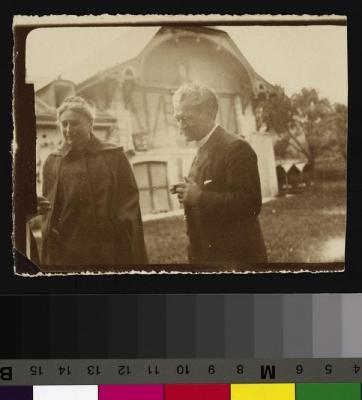 Portrait du Prof. Dr. César Roux et de sa femme à Mont-la-Ville. Il est habillé en costume et cravate et fume un cigare. Sa femme a les cheveux attachés en chignon et porte une sorte de manteau-cape. A l'arrière-plan, à gauche, une maison à colombages;…