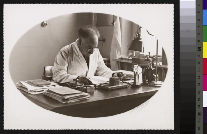 Portrait du Dr. Alfred Rosselet, assis à sa table de travail, laquelle est recouverte d'objets divers: feuilles, presse papier, papiers, lampe, téléphone. Le médecin a une cigarette dans la main gauche. A droite de l'image, une fenêtre avec un rebord s…