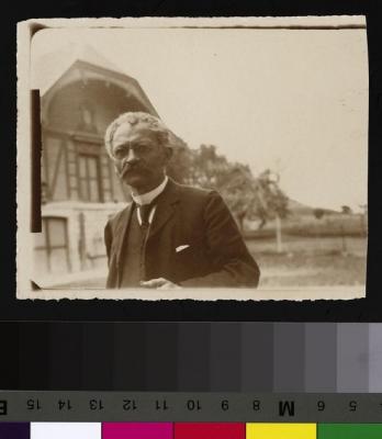 Portrait du Prof. Dr. César Roux, à Mont-la-Ville, en costume et cravate. A l'arrière-plan, à gauche, une maison à colombages; à droite, des arbres et un champ.