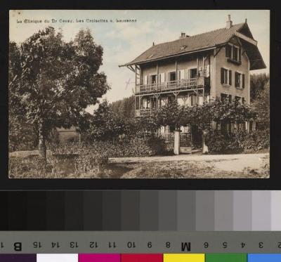 Vue sur l'extérieur de la Clinique du Dr Cevey, située aux Croisettes à Epalinges. Devant la maison, le portail donnant sur le jardin arborisé lequel est bordé par une clôture. A l'arrière plan, des champs et la forêt. Un écriteau est accroché à l'angl…