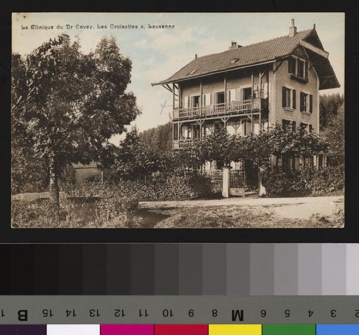 Vue sur l'extérieur de la Clinique du Dr Cevey, située aux Croisettes à Epalinges. Devant la maison, le portail donnant sur le jardin arborisé lequel est bordé par une clôture. A l'arrière plan, des champs et la forêt. Un écriteau est accroché à l'angl…
